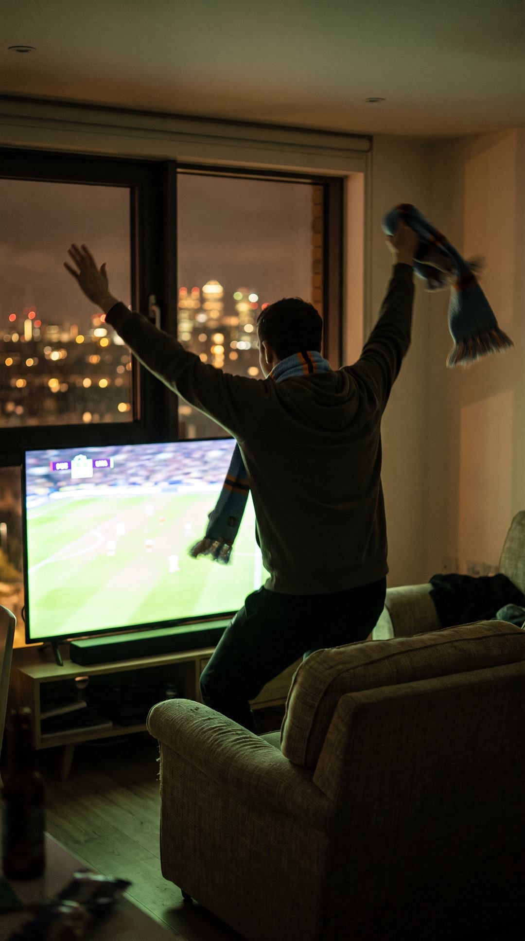 A supporter leaping out of an armchair in a dark flat at night, arms up and a scarf flying, a match on the screen and a city skyline through the window