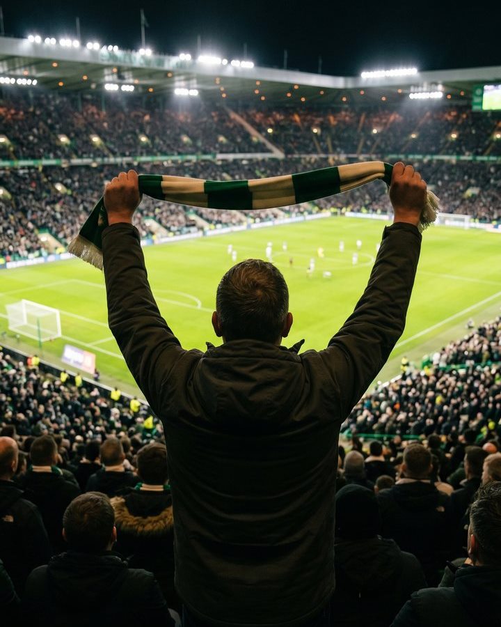 A supporter holding a green and cream scarf above his head in a packed, floodlit stadium